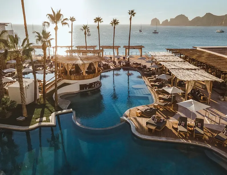 La alberca de un club de playa entre palmeras en Cabo San Lucas, con el Finisterra al otro lado de la bahía.