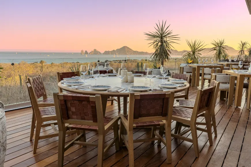 Una mesa puesta para cenar en la terraza de un restaurante al atardecer, con vista a la bahía y al Finisterra en Los Cabos.