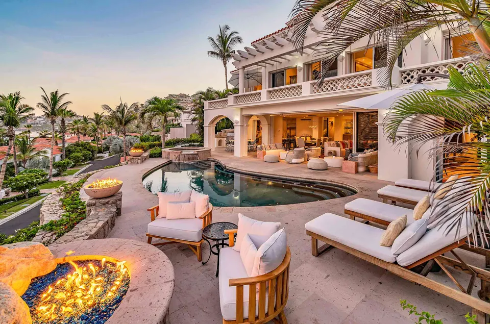 Una villa blanca en la ladera de Los Cabos al anochecer, con alberca, fogatas y camastros bajo las palmeras.